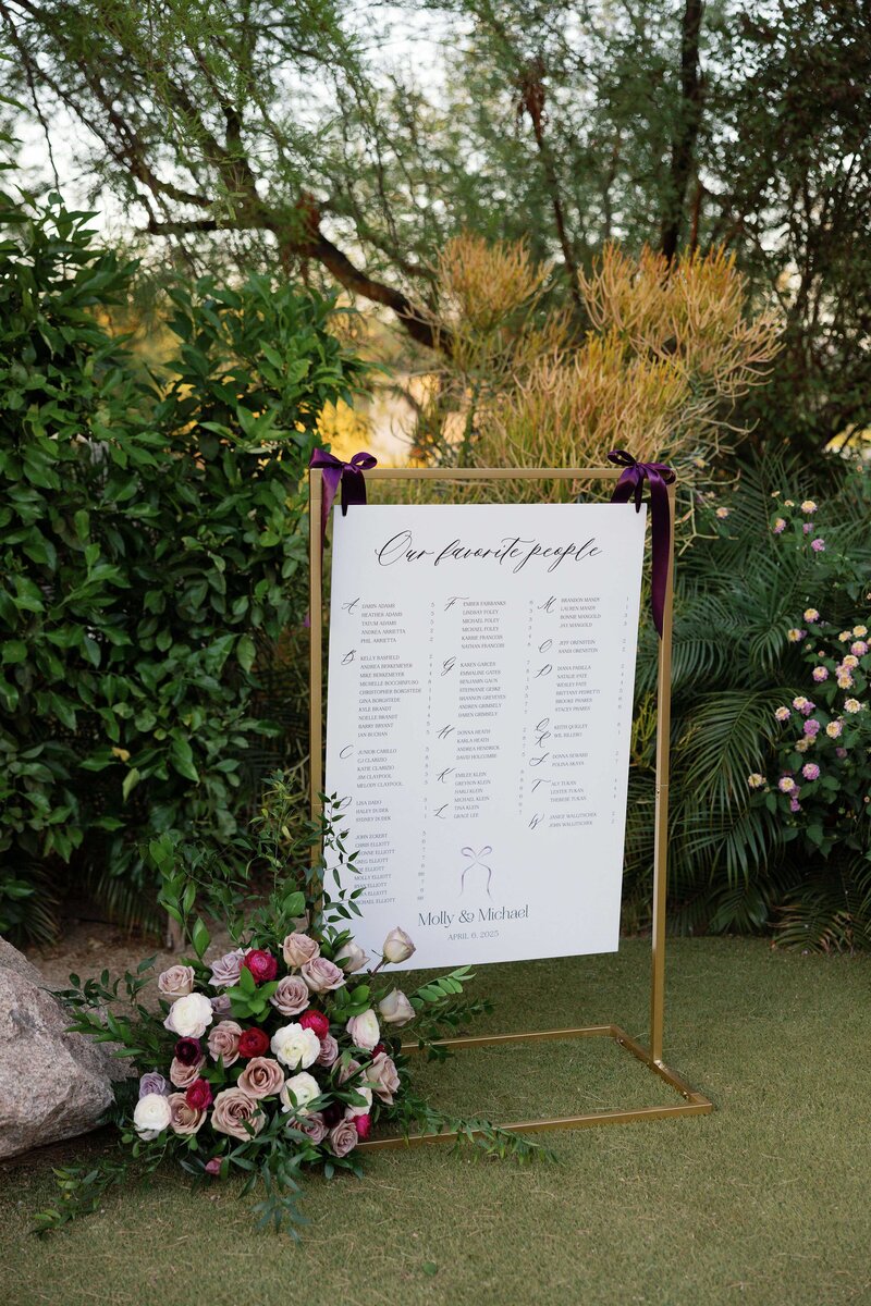 Wedding seating chart displayed on gold frame with jewel toned floral arrangement.