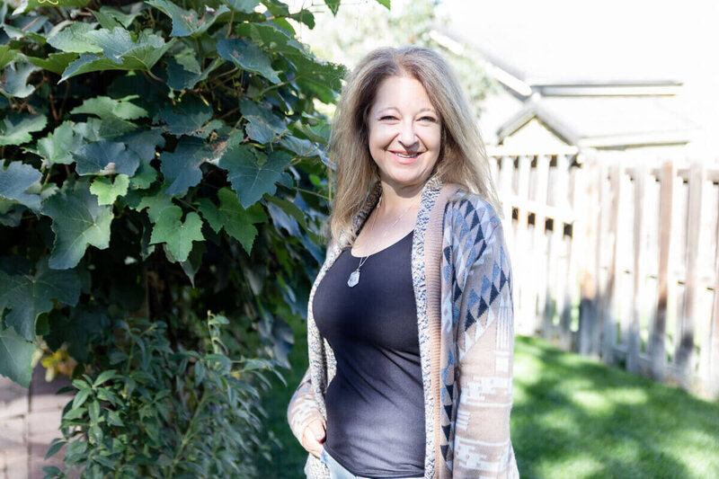 Cyndi Stewart stands in a sunlit garden next to green vines, smiling warmly.