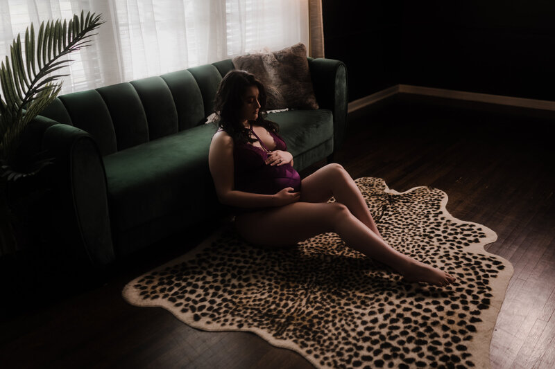A pregnant woman in purple lace lingerie sits against a couch under a window holding her bump