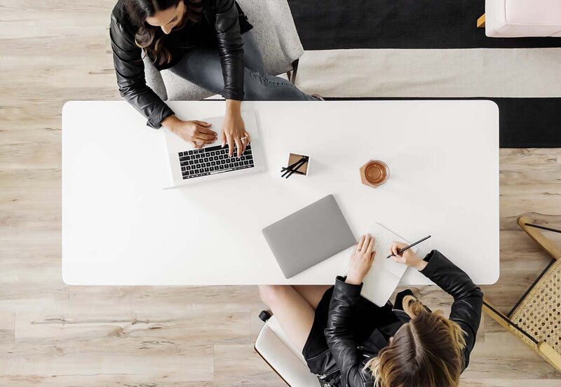 Overhead view of two women entrepreneurs working on laptops across a white table in a modern office.