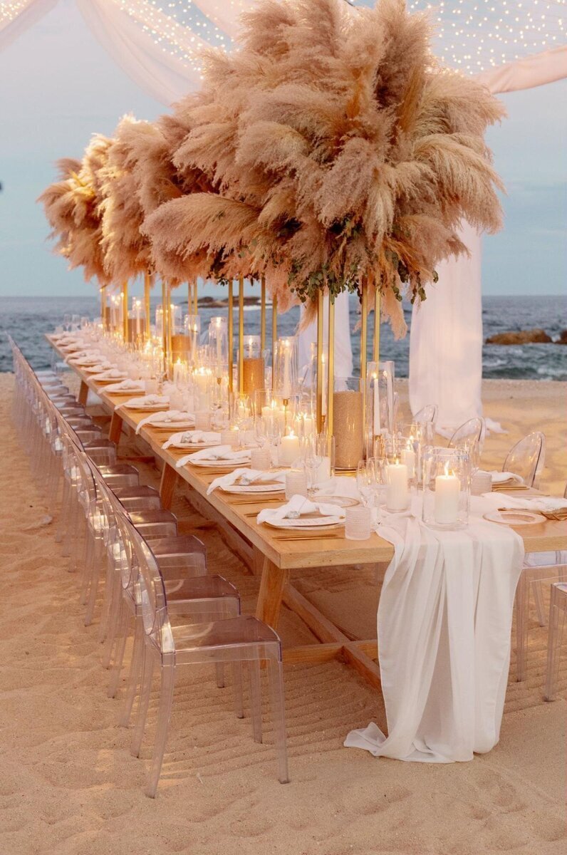 A luxurious Los Cabos wedding reception table on the beach beneath a sheer white canopy adorned with lights, a long white table runner, clear chairs, and tall pompas centerpieces
