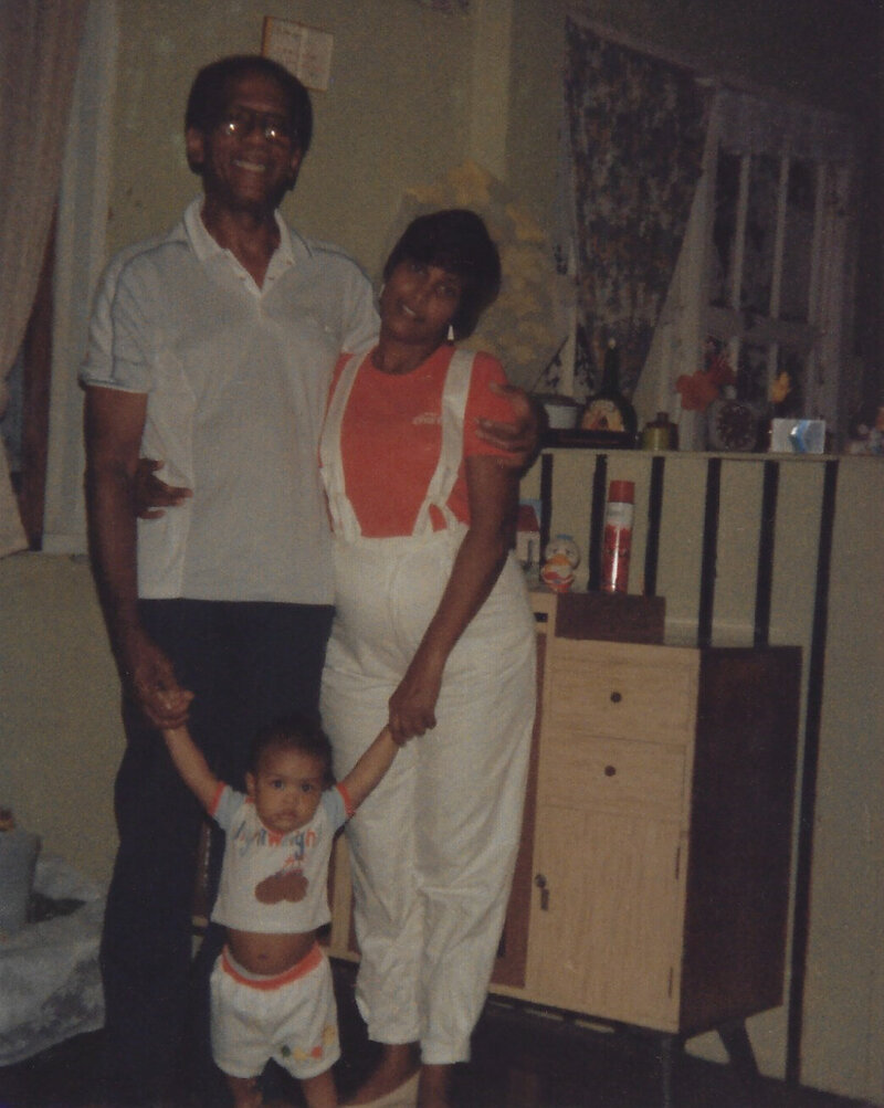 A vintage family photo taken in the 1980s in Guyana, South America, showing my mother and father each holding one of my hands as I stand between them as a baby, captured inside our home, part of our family's legacy.