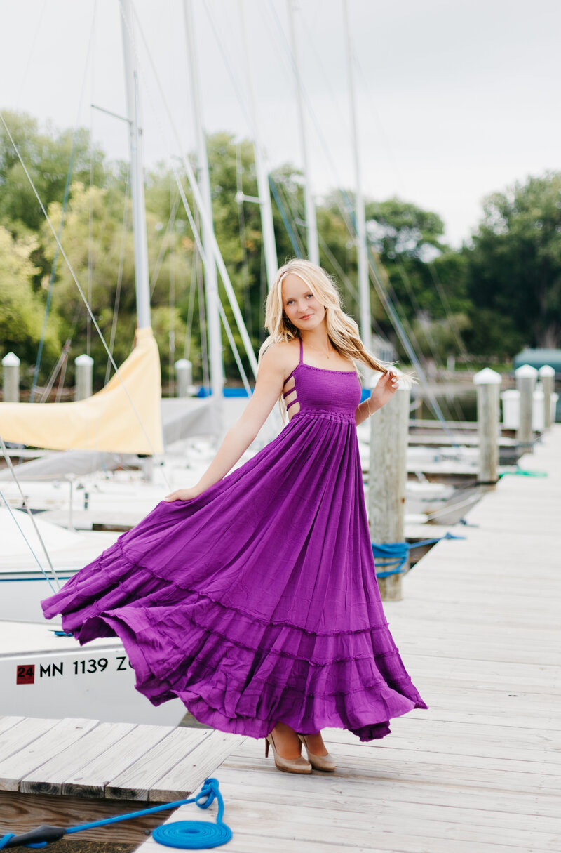 Senior girl in a long purple dress holding her hair and twirling her dress on a dock in front of sailboats