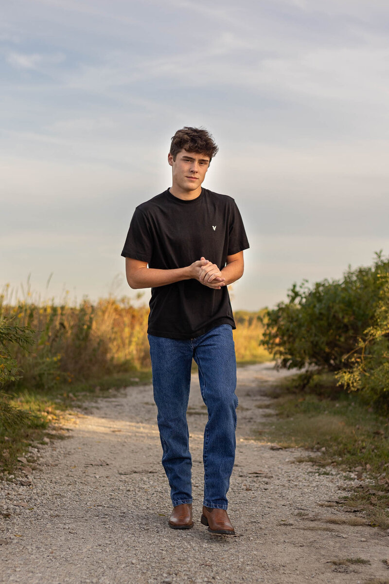 A senior boy walks along a gravel path at sunset, dressed in a classic black t-shirt and jeans with brown boots. The soft golden light highlights his confident and relaxed demeanor, making this a timeless and natural outdoor senior portrait.