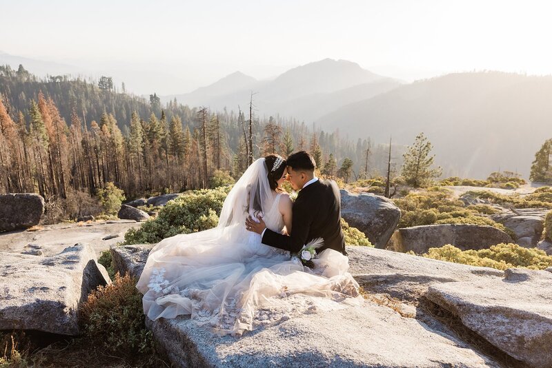 An elopement bride and groom sit on the mountain top and look at each other during sunset.