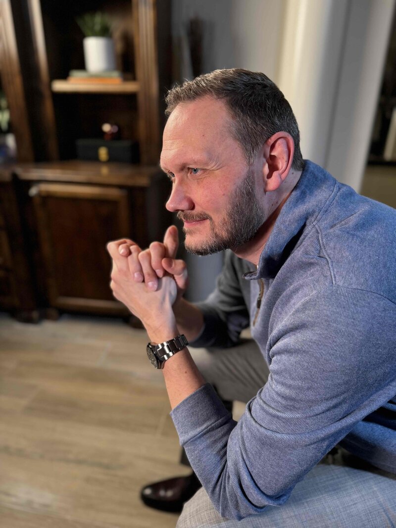 middle-aged man leaning forward in a chair with hands clasped and elbows resting on his knees. He is intently listening which promotes a feeling of caring and sincerity