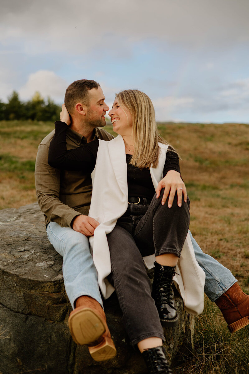 a couple sitting on a rock embracing with a rainbow in the background during their engagement session in Connecticut
