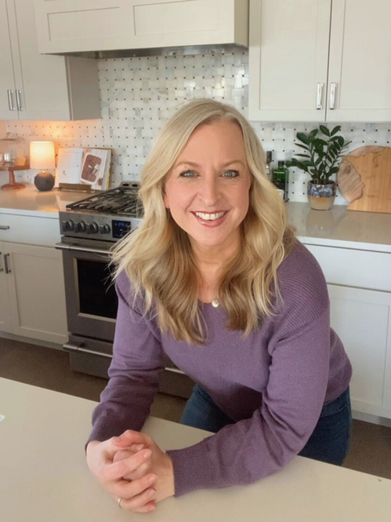 Woman with blonde hair wearing a purple sweater, smiling and leaning on a kitchen counter.