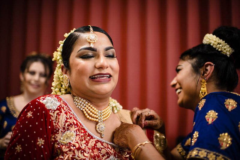 Bride in traditional hindu wedding attire smiles at the camera while her sister fixes her dress at a CT indian wedding