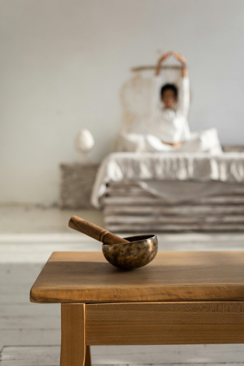 focus is on metal singing bowl sat upon wooden tabletop, background is a woman with legs crossed, dressed in white clothing, reaching up to stretch while sitting on her bed