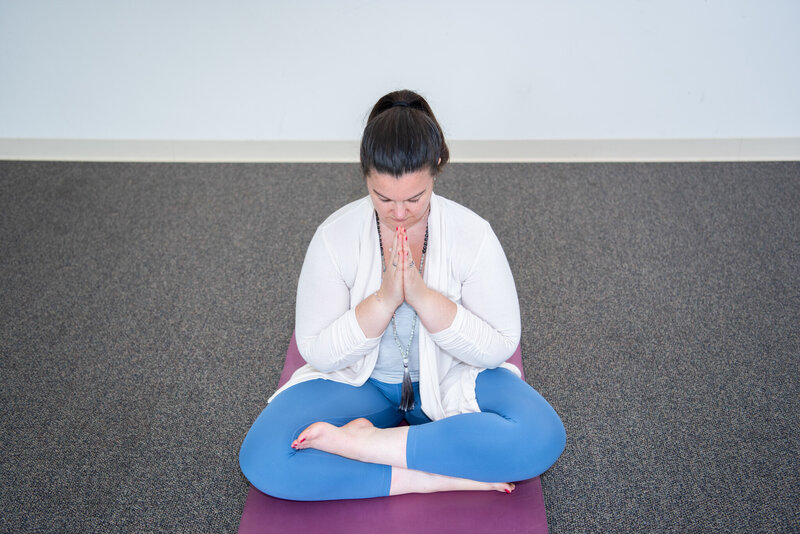 A woman sitting on her yoga mat with her legs crossed and hands together in front of her chest.