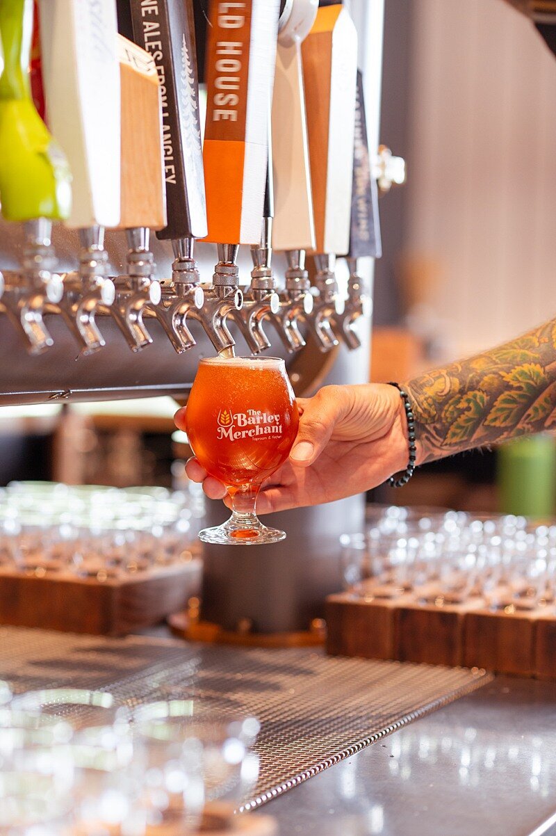 hand holding a full beer glass with the Barley Merchant logo on it under a tap