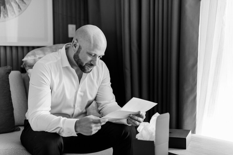 A bald man with a beard, dressed in a white dress shirt, sits on a couch reading a letter. A gift box and tissue paper are on the seat next to him. He is inside a room with a curtain drawn to let light in from the window.