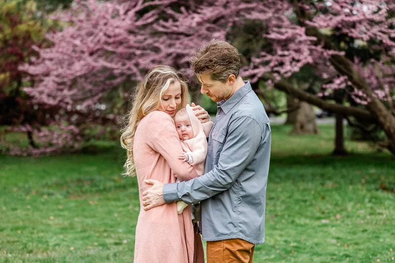 Mother holding baby and man embracing family in front of a cherry blossom tree