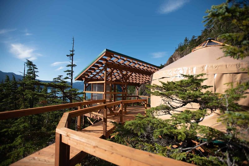 A light colored, luxury glamping yurt  and an expansive outdoor deck and lounging space on a platform overlooking Resurrection Bay in Alaska during a luxury Kenai Fjords and glacier elopement .  There is a beautiful blue sky, and the lush green of the fjords at Shearwater  Cove Glamping surrounds the yurt with the dark blue of the water in the bay in the distance. 