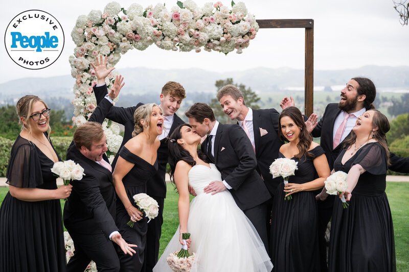 A newlywed couple shares a romantic kiss under a breathtaking floral arch at Spanish Hills, surrounded by their excited bridal party in elegant black attire. The candid moment captures pure joy and celebration, beautifully documented by Los Angeles wedding photographers.