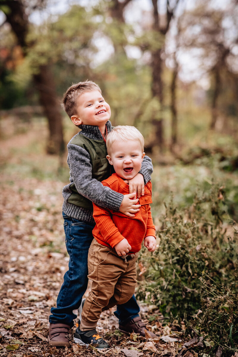 Two young boys, one in a green vest and the other in an orange sweater, embrace each other with big smiles while standing on a leaf-covered path in a wooded area.
