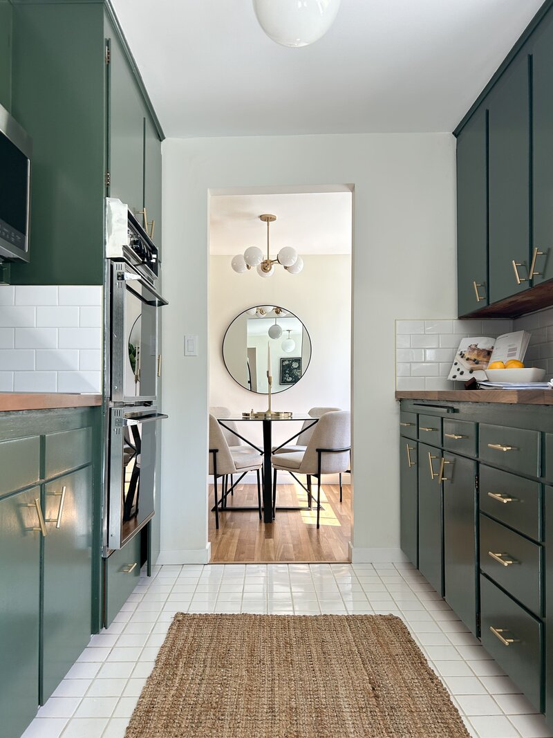 Sage green kitchen styled by Modern Mollusk with a jute rug, modern kitchen decor, and a round framed mirror.