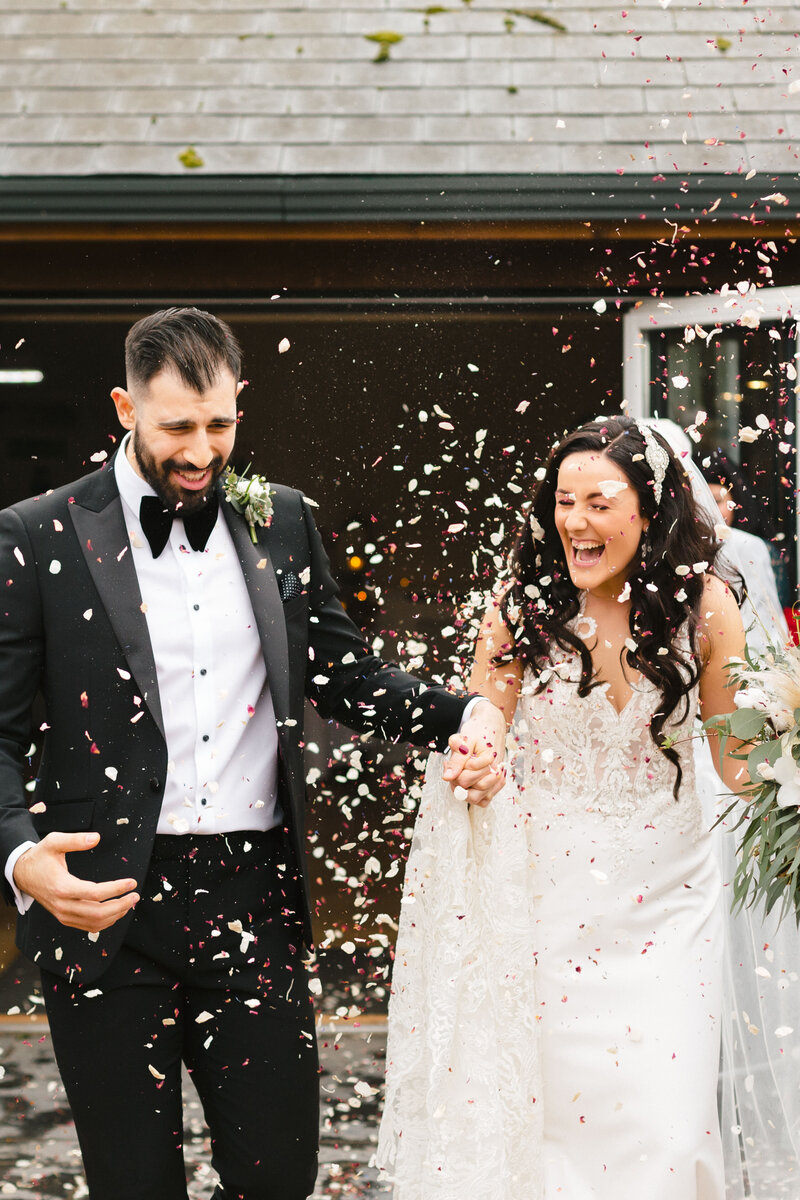 A bride and groom joyfully walk through a shower of confetti at Wasing Park, surrounded by cheering guests. The bride wears a white gown and veil, holding a bouquet, while the groom is in a black suit. The scene is festive and celebratory.