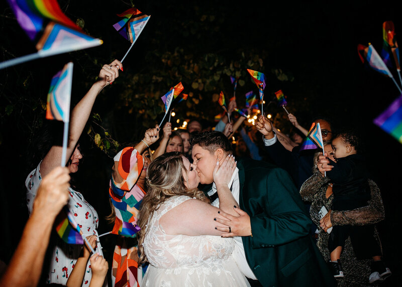 A newlywed couple kissing while guests wave pride flags around them.