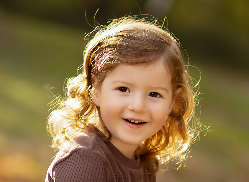 Little boy's brooklyn upsherin photoshoot. The little boy has soft golden light highlighting the curls in his hair. He is looking over his shoulder and smiling at the camera. Captured by top Brooklyn, NY newborn photographer Chaya Bornstein Photography.