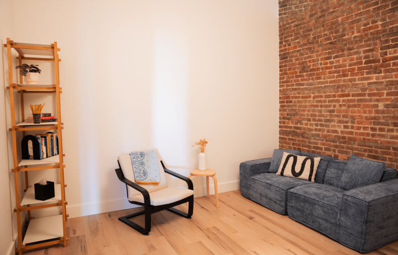 Office with cozy blue sofa, abstract pillows and a chair with a small side table and flowers atop.