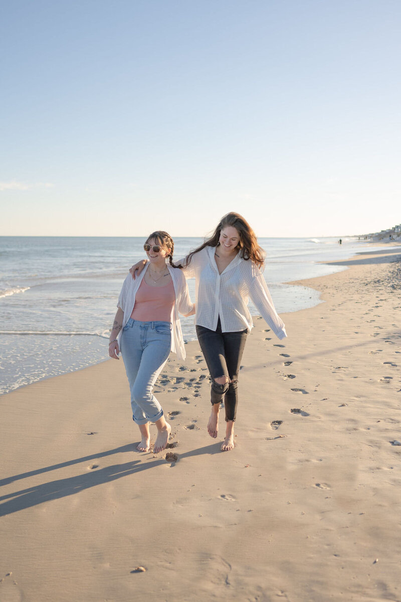 two women walk barefoot on the beach with waves and footprints in the background