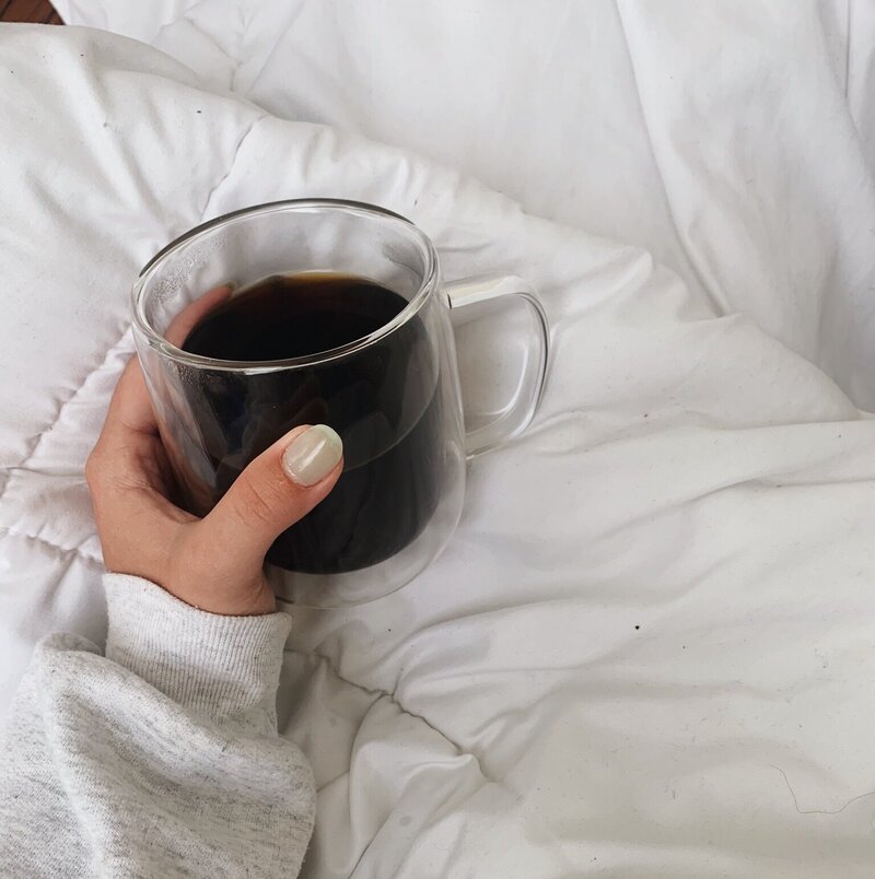 A hand holding a clear mug of black coffee rests on white bedding. The person wears a gray sweater, creating a cozy and relaxed atmosphere.