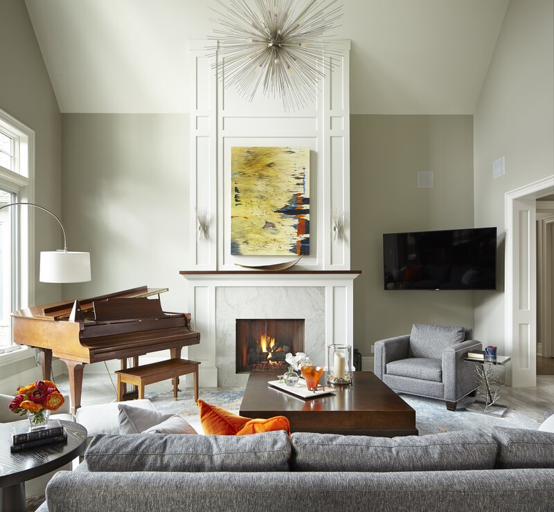 Living room design, the room has light sage green walls, a grey sofa and matching chair, ornage pillows, a wooden baby brand piano, and white trimmed and marble fireplace on the wall centre