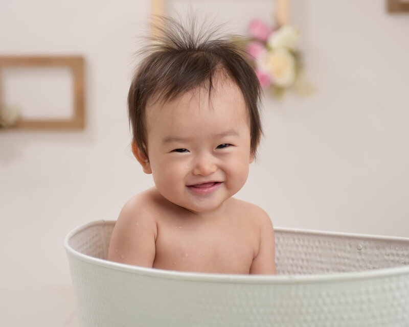 A baby girl wearing a floral pink headband, sitting in a white tub with pink and white balloons in the background.