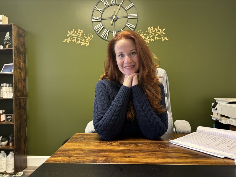 Jenn smiling at her desk with a green wall and decorative clock in the background, surrounded by office supplies and notes