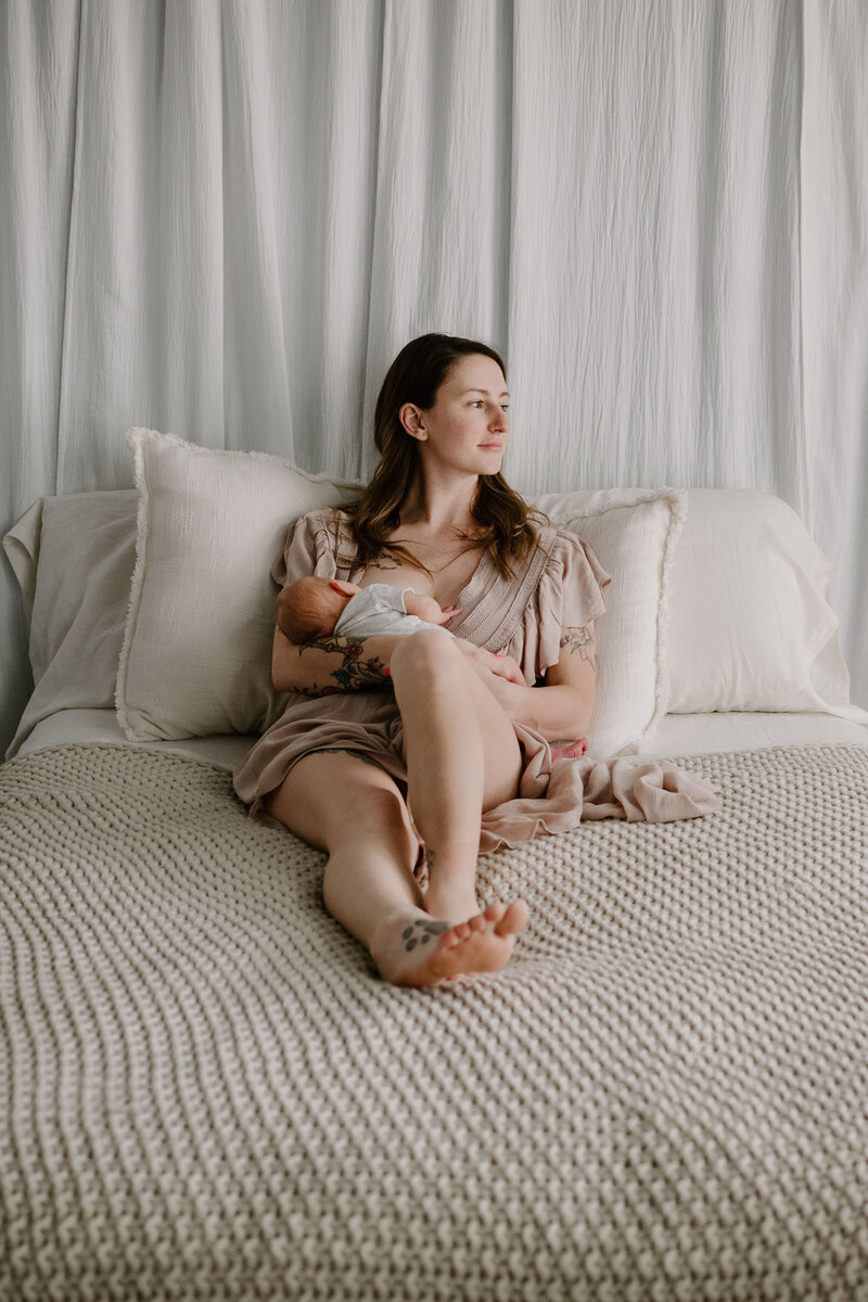 Mom in tan holding baby while sitting on white bed in studio in Severn Maryland photographed by Bethany Simms Photography