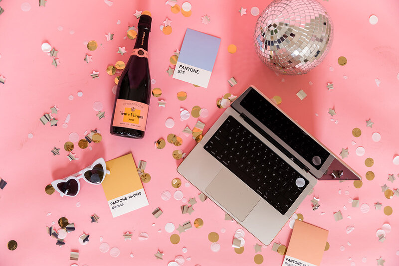 laptop, champagne, and pantone swatches laying on pink background with confetti