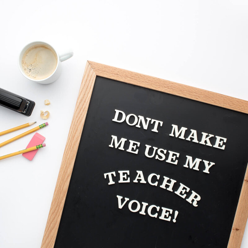 chalk board that says don't make me use my teacher voice next to a cup of coffee with milk, a black stapler, three yellow number 2 pencils, and a pink eraser