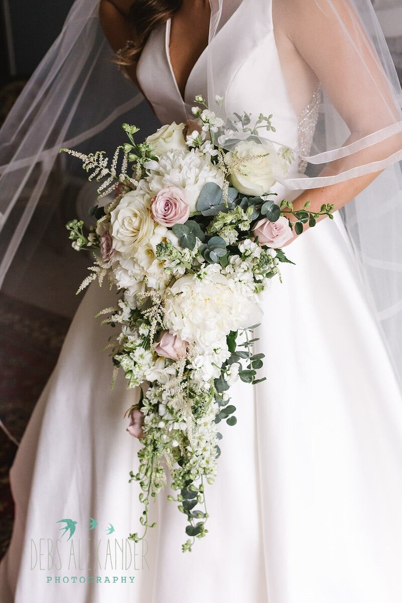 A bride in a white stylish dress carrying a cascading bouquet of white and blush flowers for her wedding in Cornwall