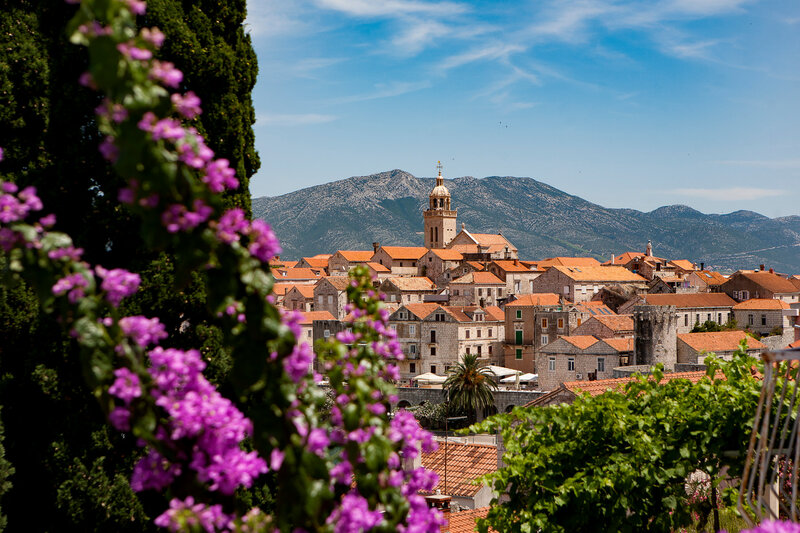 Explore this charming historic town, a gem for any European travel specialist, with its terracotta rooftops and central church tower nestled against a stunning mountainous backdrop. In the foreground, vibrant purple flowers and lush greenery bask under the radiant blue sky.