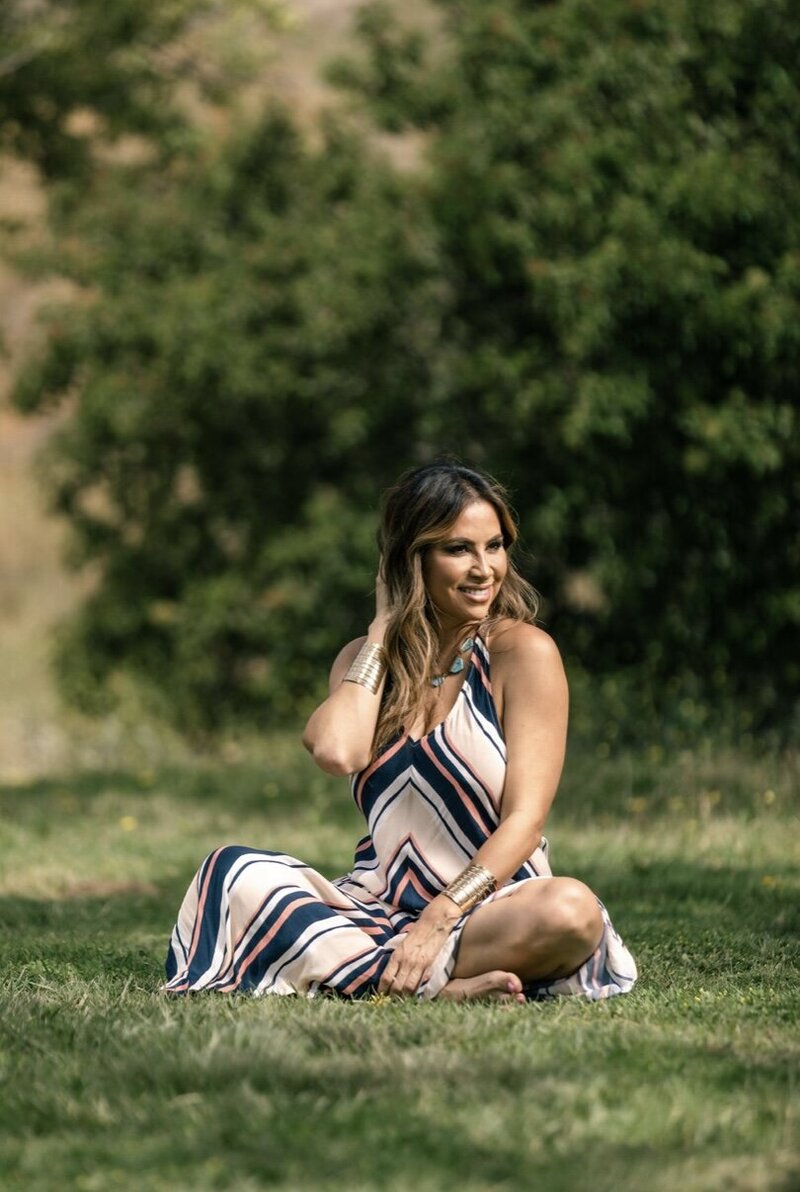 Mayra Menjivar, Warrior Woman Founder, sitting in the grass in a striped dress