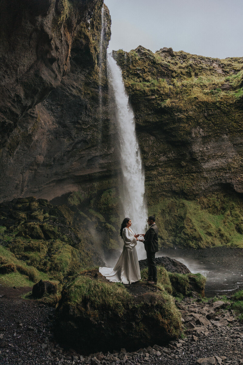 Couple holds hands on a rock near an Icelandic waterfull