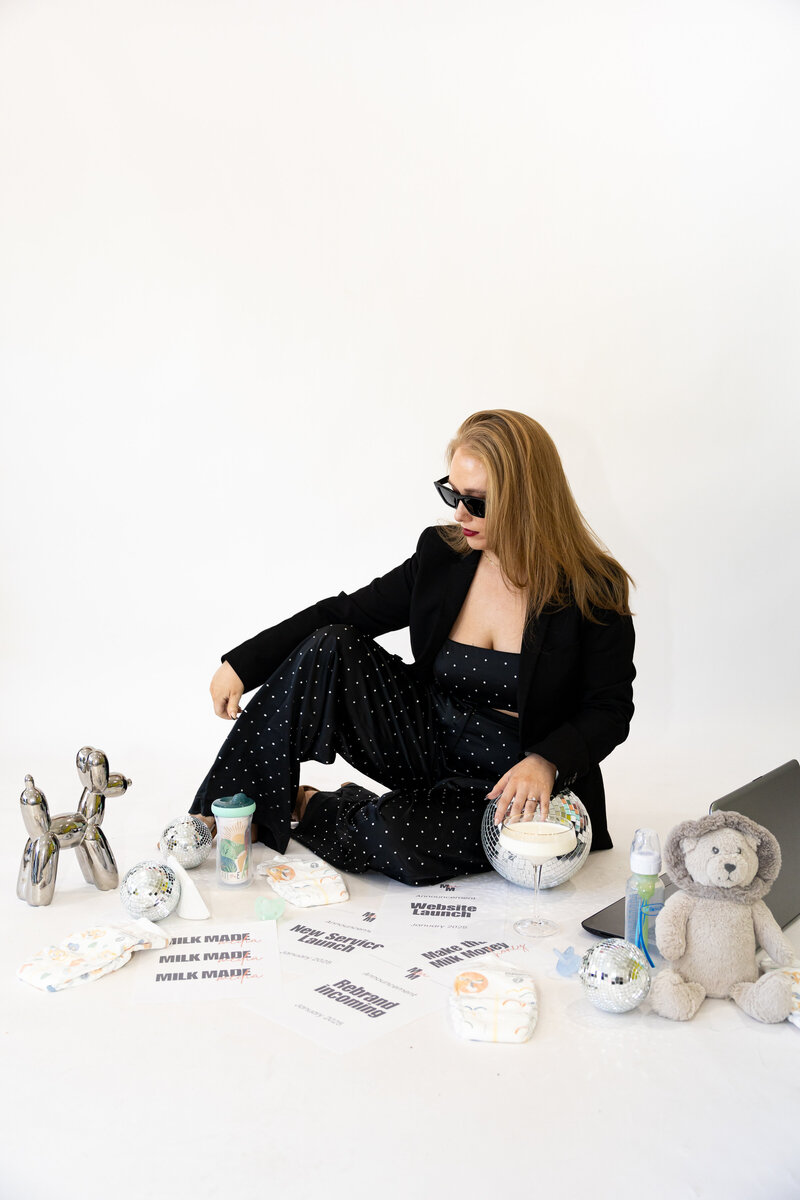 photo of a caucasian woman wearing sunglasses sitting on the floor surrounded by pieces of paper, disco  balls, diapers, a baby monitor, a laptop, a teddy bear, and a sippy cup