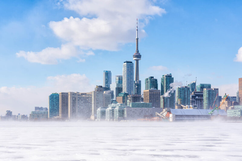 Winter view of downtown Toronto skyline with the CN Tower and modern high-rise buildings, seen across a frozen Lake Ontario under a bright blue sky with scattered clouds.