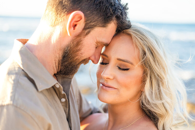 man and woman close up of their faces nuzzling and ocean in the background