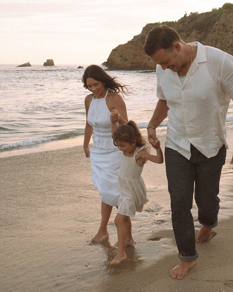Parents holding their daughter’s hands while walking along the shore at Laguna Beach, playful and candid at golden hour.