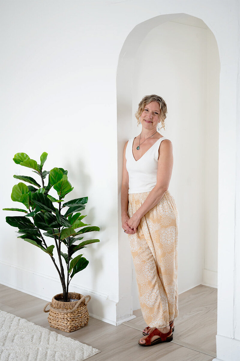 Heidi Schauster stands in front of a white wall, next to a vibrant potted plant, smiling warmly at the camera.