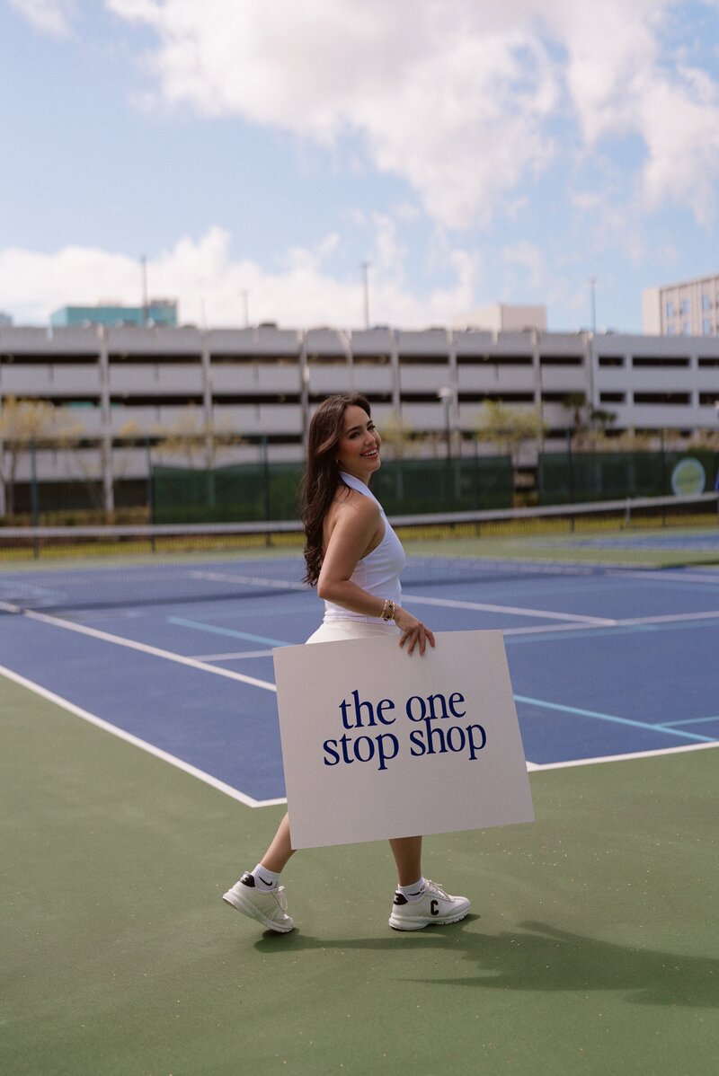 Woman on tennis court holding sign saying the one stop shop