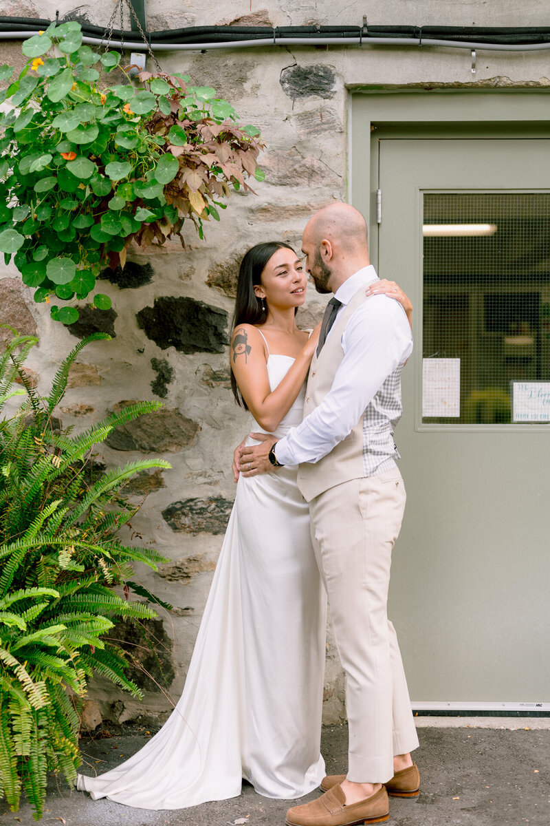 A couple embracing and smiling at each other in front of a stone wall with greenery.