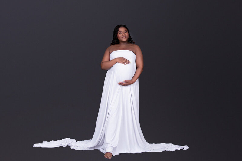 Mom-to-be in a white maternity gown posing against a dark studio backdrop