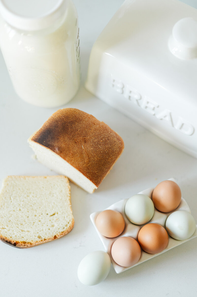 Bright styled photo of homemade bread, multi-colored eggs, and milk on a white kitchen counter.