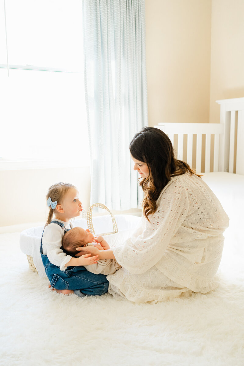 Austin Texas Portrait Photographer | A mother and her two children sit on the floor in a nursery. The mother places her newborn baby into her daughter's lap. The daughter talks to her mother.