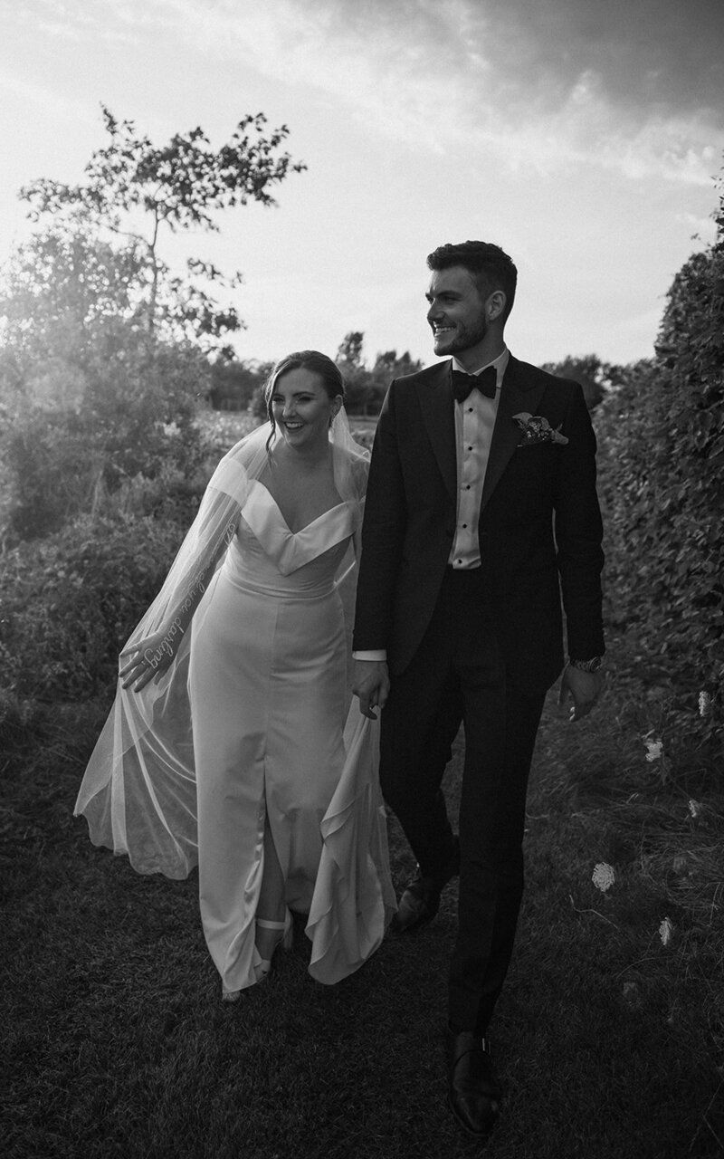 Black and white image of a joyful bride and groom walking hand in hand outdoors, with the bride in an elegant gown and veil, and the groom in a classic tuxedo, surrounded by lush greenery and a soft, evening light.