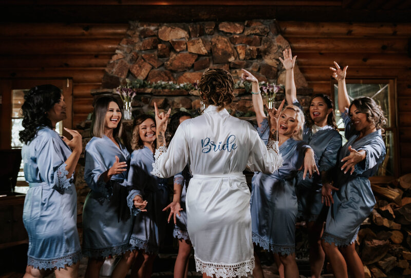 Bride throwing up rockstar hands in custom robe while her bridal party hypes her up in the bridal suite